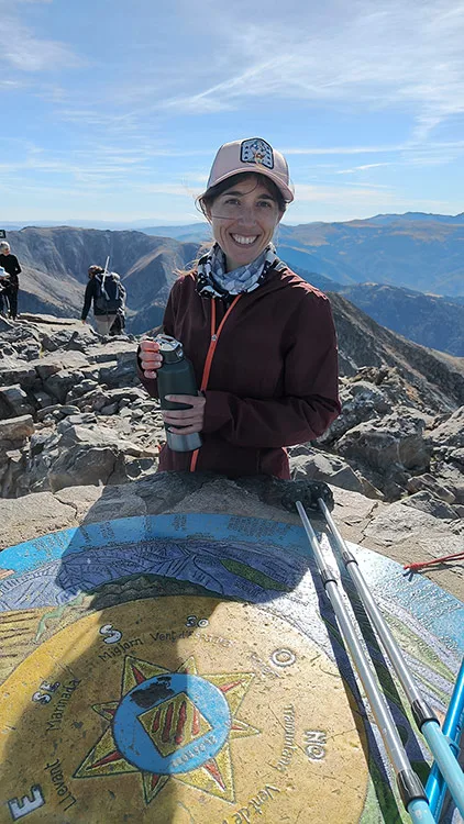 Emilie tient une gourde devant la table d'orientation située au pic du Canigou